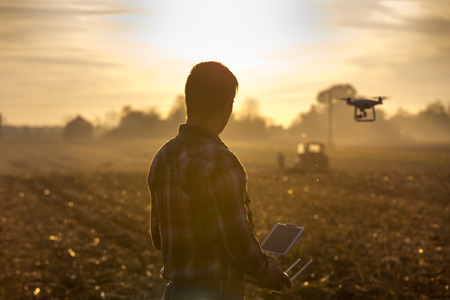Attractive farmer navigating drone above farmland with silos and tractor in background. High technology innovations for increasing productivity in agricultureの写真素材