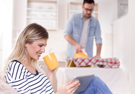 Beautiful young woman resting on sofa and looking at tablet while man ironing her shirt in background. Husband doing chores conceptの写真素材