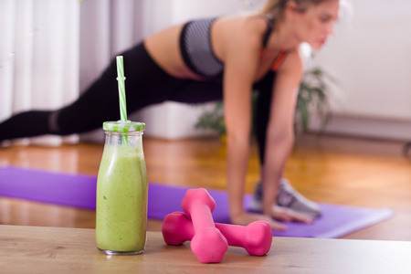 Close up of dumbbells and smoothie in jar with straw and pretty young woman stretching in...