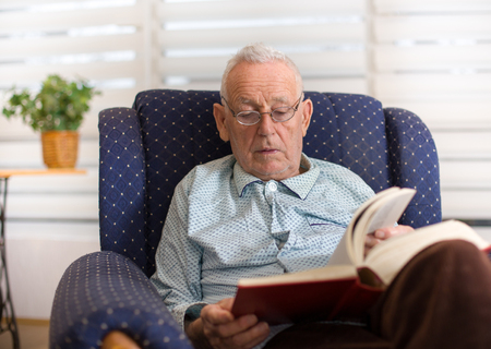 Portrait of serious old man reading book in armchair at homeの写真素材