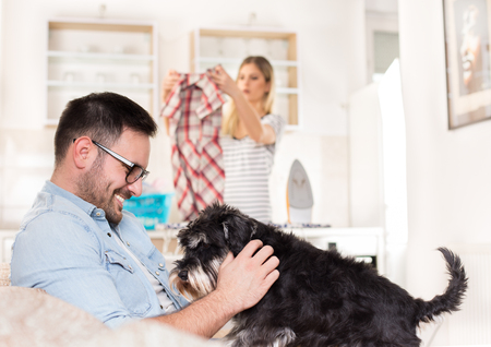 Young handsome man sitting on sofa and cuddling dog while wife ironing and doing chores in backgroundの写真素材