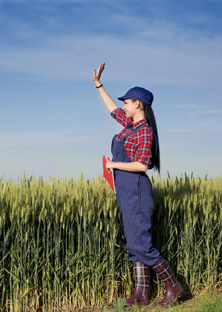 Pretty young farmer woman standing in front of green wheat field and waving handの写真素材