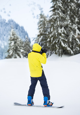 Young man on snowboard with photo camera taking shoot on ski slope. Winter sports and vacation conceptの写真素材