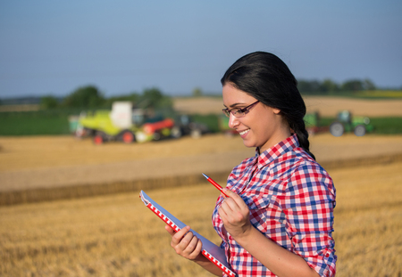 Pretty young woman with note book standing on wheat field during harvestの写真素材