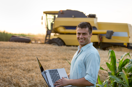 Happy young farmer engineer with laptop standing on wheat field while combine harvester working in...