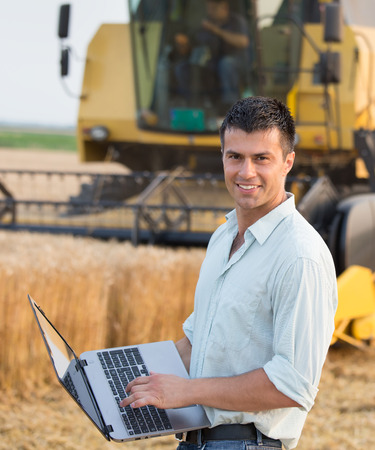 Happy young farmer engineer with laptop standing on wheat field while combine harvester working in backgroundの写真素材