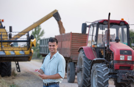 Satisfied young farmer with notebook standing in front of combine harvester while loading trailer...