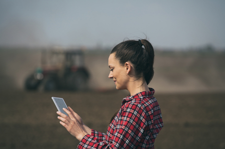 Pretty young woman with tablet standing in front of tractor in fieldの写真素材