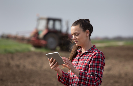 Pretty young woman with tablet standing in front of tractor in fieldの写真素材