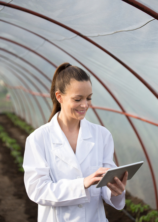 Young pretty woman agronomist in white coat with tablet supervising seedlings in greenhouse. Plant care and protection conceptの写真素材