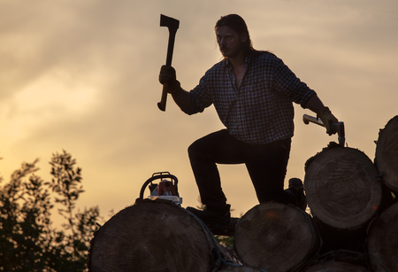 Strong lumberjack swinging with ax above wooden trunk in forest at sunsetの写真素材