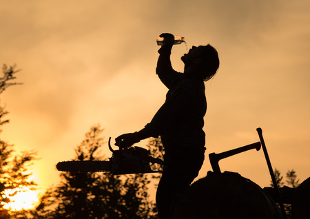 Silhouette of lumberjack with chainsaw drinking water from bottle on wooden log pile at sunset in hot summer eveningの写真素材