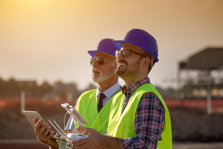 Two engineers with helmets and vests operating with drone by remote control and looking up in the...