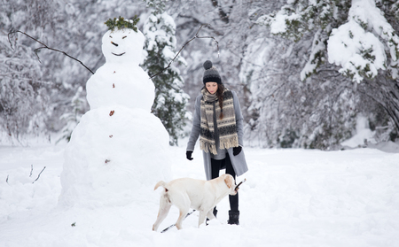 Young woman building snowman with help from her cute dog with branch in mouth in parkの写真素材