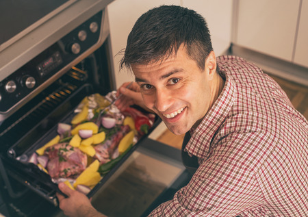 Satisfied young man putting casserole with meat and vegetables in oven