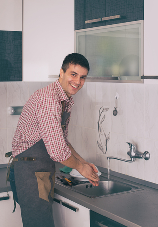 Young handsome man with apron washing dishes in kitchen after preparing mealの写真素材