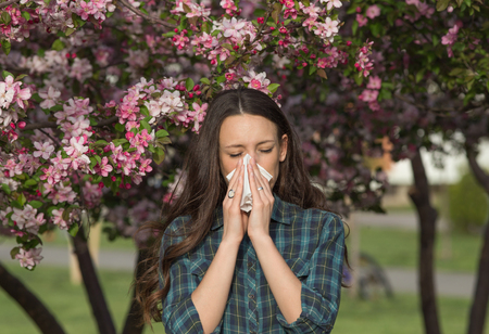 Young pretty girl blowing nose in front of blooming tree. Spring allergy conceptの写真素材