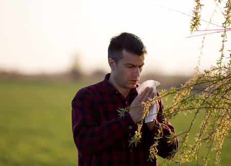 Young handsome man having allergy symptoms beside blooming tree in spring