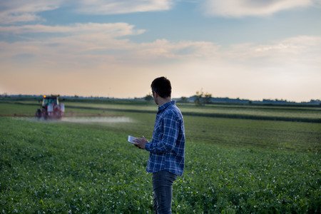 Young attractive farmer with tablet standing in front of tractor in fieldの写真素材