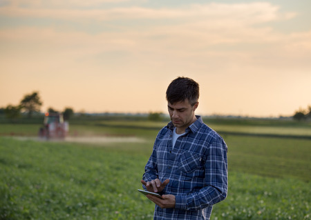 Attractive young farmer working on tablet in field with tractor spraying crops in spring
