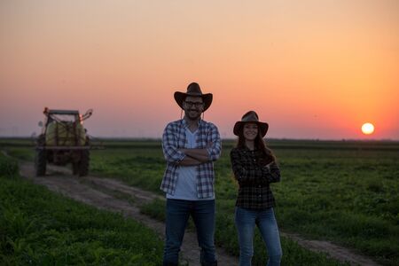 Two farmers standing with crossed arms in front of tractor at sunsetの写真素材