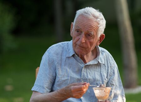 Senior man sitting in garden and eating ice cream from glass bowl, with green background