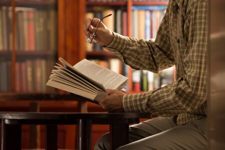 Mature man sitting at table and holding book in front of shelves in libraryの写真素材