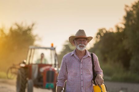 Portrait of mature farmer with straw hat holding plant sprayer in front of tractor in field at sunsetの写真素材