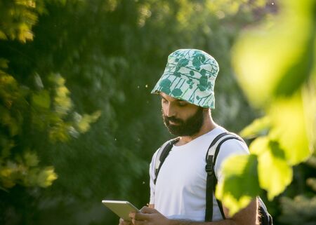 Young attractive man with beard hiking in forest with backpack on shoulders and looking at tablet