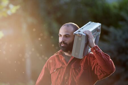 Young handsome man with beard holding vintage tape recorder on shoulder and walking in park at...