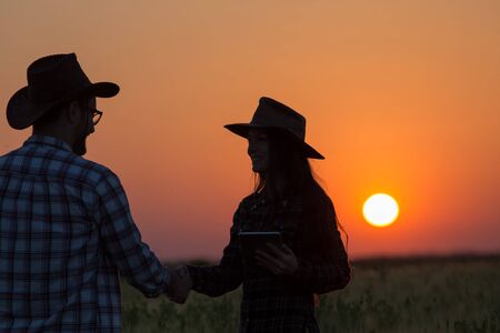 Silhouettes of man and woman farmers with hats shaking hands in field during sunsetの写真素材