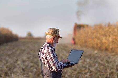 Senior farmer with laptop standing in front of combine harvester in corn field in autumnの写真素材