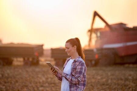 Farmer woman holding tablet in front of combine harvester loading trailer with corn grains in field at sunsetの写真素材