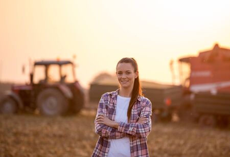 Satisfied young farmer woman standing with crossed arms in front of combine harvester and tractor with trailer in corn fieldの写真素材