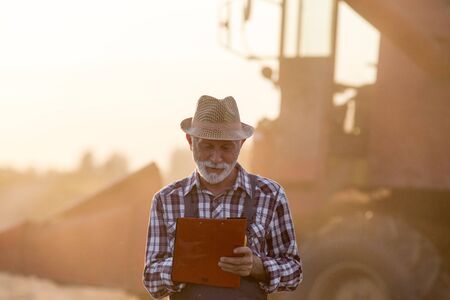 Senior man writing notes about yield in front of combine harvester at sunsetの写真素材