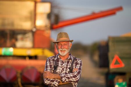 Portrait of senior farmer standing with crossed arms in front of combine harvester and tractor with...