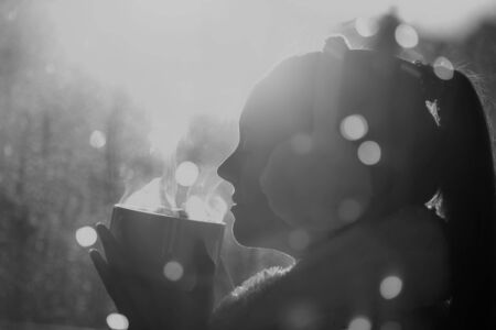 Beautiful girl covered with blanket and earmuffs holding cup of hot coffee with christmas lights at sunset. Festive moments conceptの写真素材