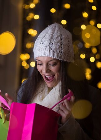 Excited young girl in winter clothes looking at shopping bags in front of shop window with christmas lights in backgroundの写真素材