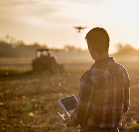 Attractive farmer navigating drone above farmland with tractor in background. High technology...