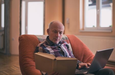 Mature man sitting in armchair and doing research on laptop and in bookの写真素材