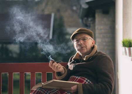 Senior man sitting on bench in garden covered with blanket reading book and smoking pipeの写真素材