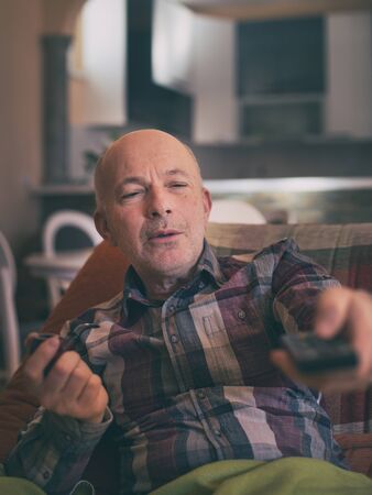 Mature man sitting on sofa at home holding remote control, smoking pipe and watching TVの写真素材