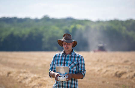 Farmer with hat holding remote control of drone and supervising harvest in golden wheat field