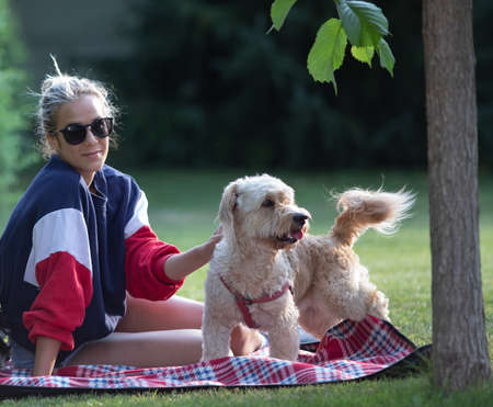 Pretty young girl sitting on blanket in park and pet her cute dog
