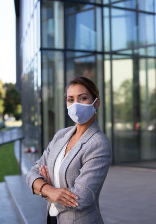 Beautiful young business woman standing with crossed arms and wearing facial protective mask in front of glass building. Pandemic protection and responsibility in businessの写真素材