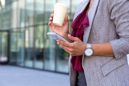 Close up of business woman's hands holding cup of coffee and typing massage on mobile phone, in front of glass office buildingの写真素材
