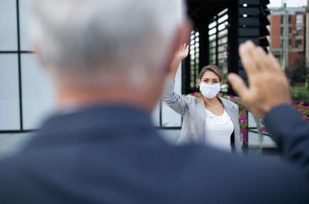 Business woman with safety mask and man greeting by waving hands in front of office building. Virus protection and social distancing concept at workの写真素材