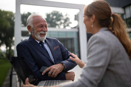Young business woman talking to mature man in suit in front of office building, laughing and holding laptop on terraceの写真素材