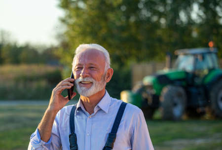 Portrait of satisfied senior farmer talking on mobile phone in front of tractor in background on farmの写真素材