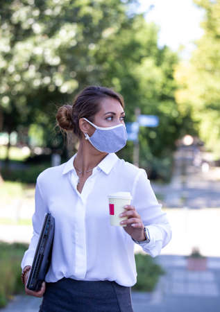 Attractive business woman with facial mask carrying laptop and cup of coffee outdoorの写真素材